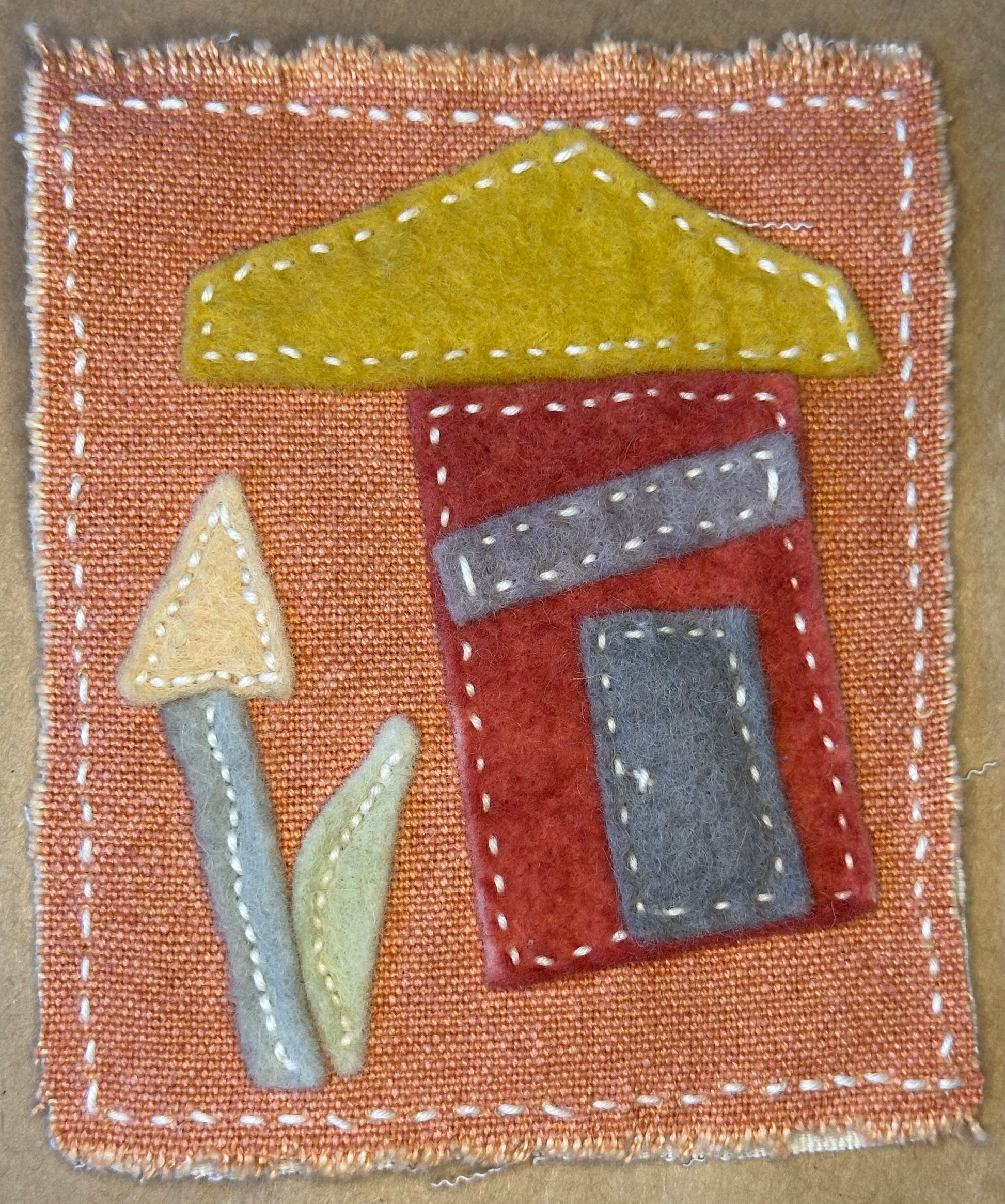 Textile artwork by Suzanne Gibbs from her Fragments of Home series, featuring a red house with a golden roof and an abstract flower on a cinnamon-colored fabric background.