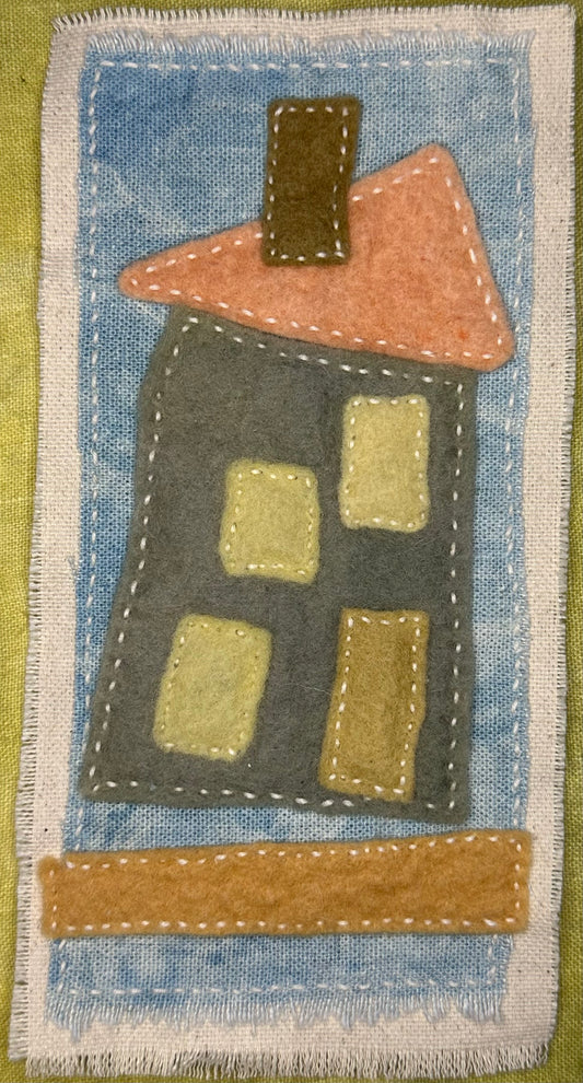 Fragments of Home textile art by Suzanne Gibbs with a gray felt house, peach roof, green chimney, and yellow windows on a blue fabric background, hand-stitched in white.