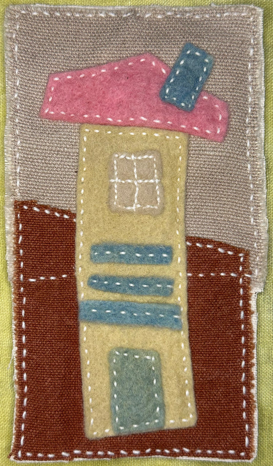 Fragments of Home textile art by Suzanne Gibbs with a tall pale yellow felt house, pink roof, blue chimney, and stitched blue details on a brown and tan fabric background, hand-stitched in white.