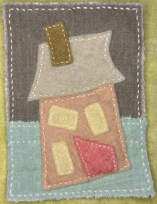 Fragments of Home textile art by Suzanne Gibbs with a tilted peach felt house, pale gray roof, olive chimney, yellow windows, and pink door on a teal and gray fabric background, hand-stitched in white.