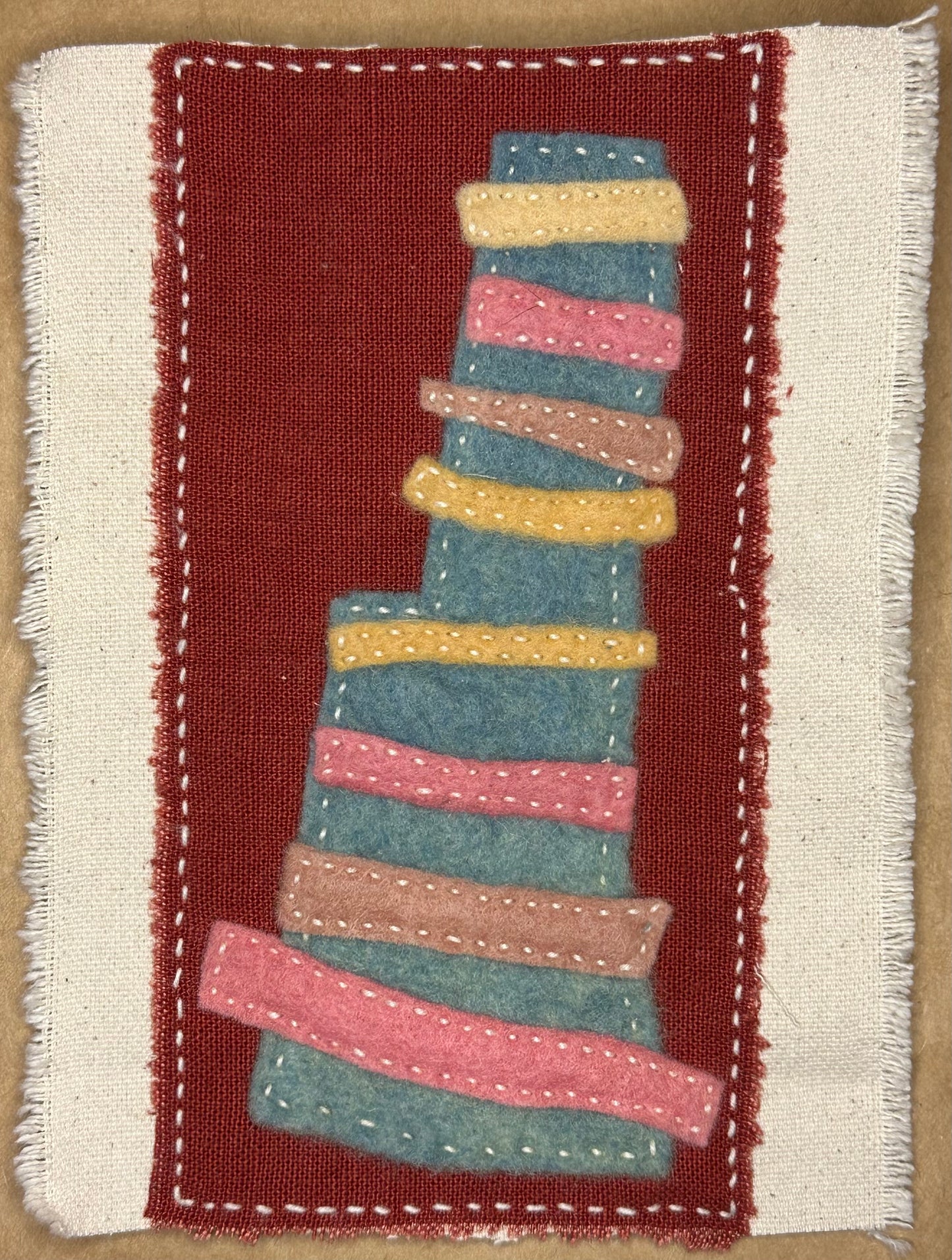 Fragments of Home textile art by Suzanne Gibbs with a leaning blue felt tower layered with pink, yellow, and tan stitched bands on a deep rust fabric background, hand-stitched in white.