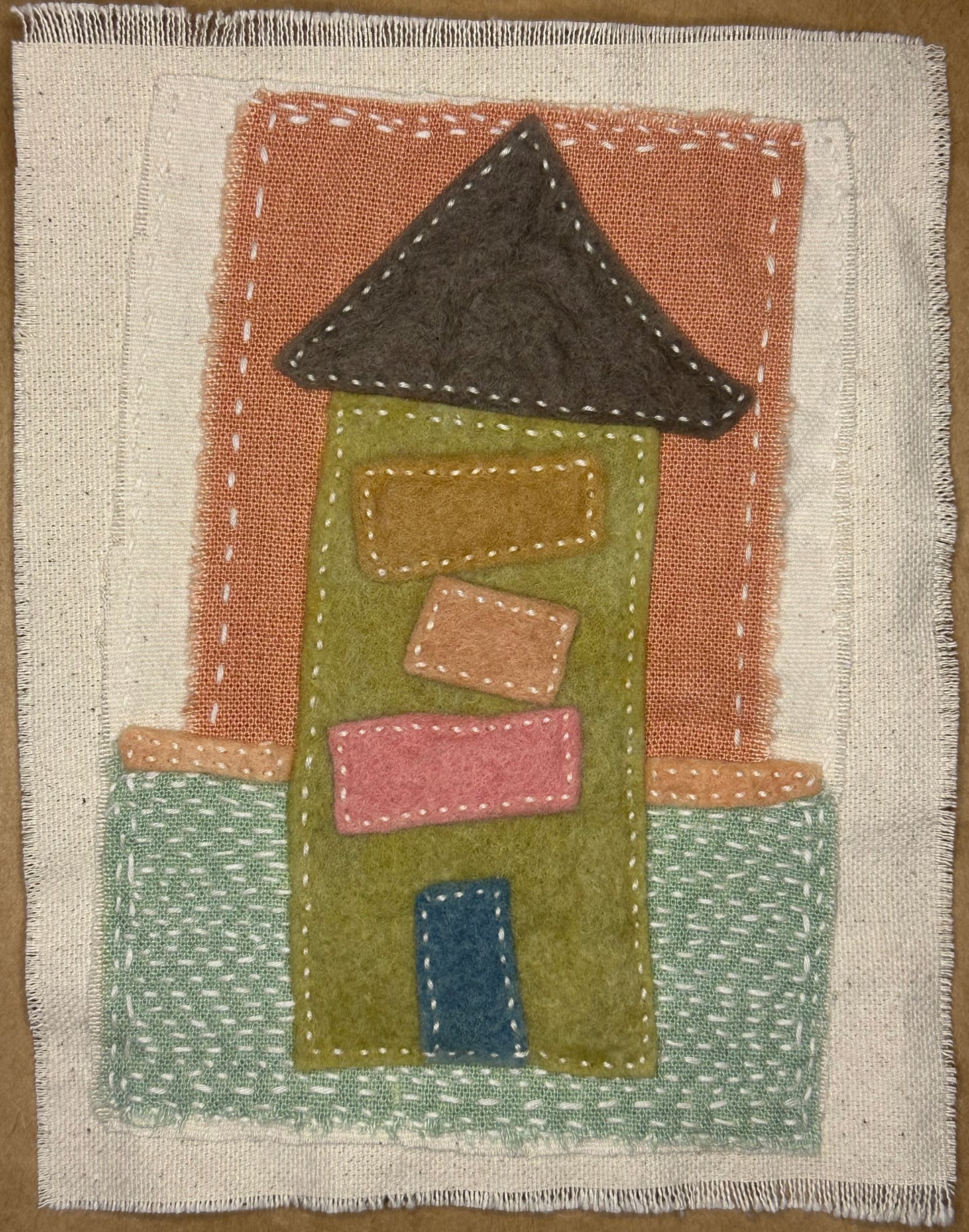 Fragments of Home textile art by Suzanne Gibbs with a green felt house, dark brown roof, pink and tan windows, blue door, and a base resembling mowed grass, hand-stitched in white.