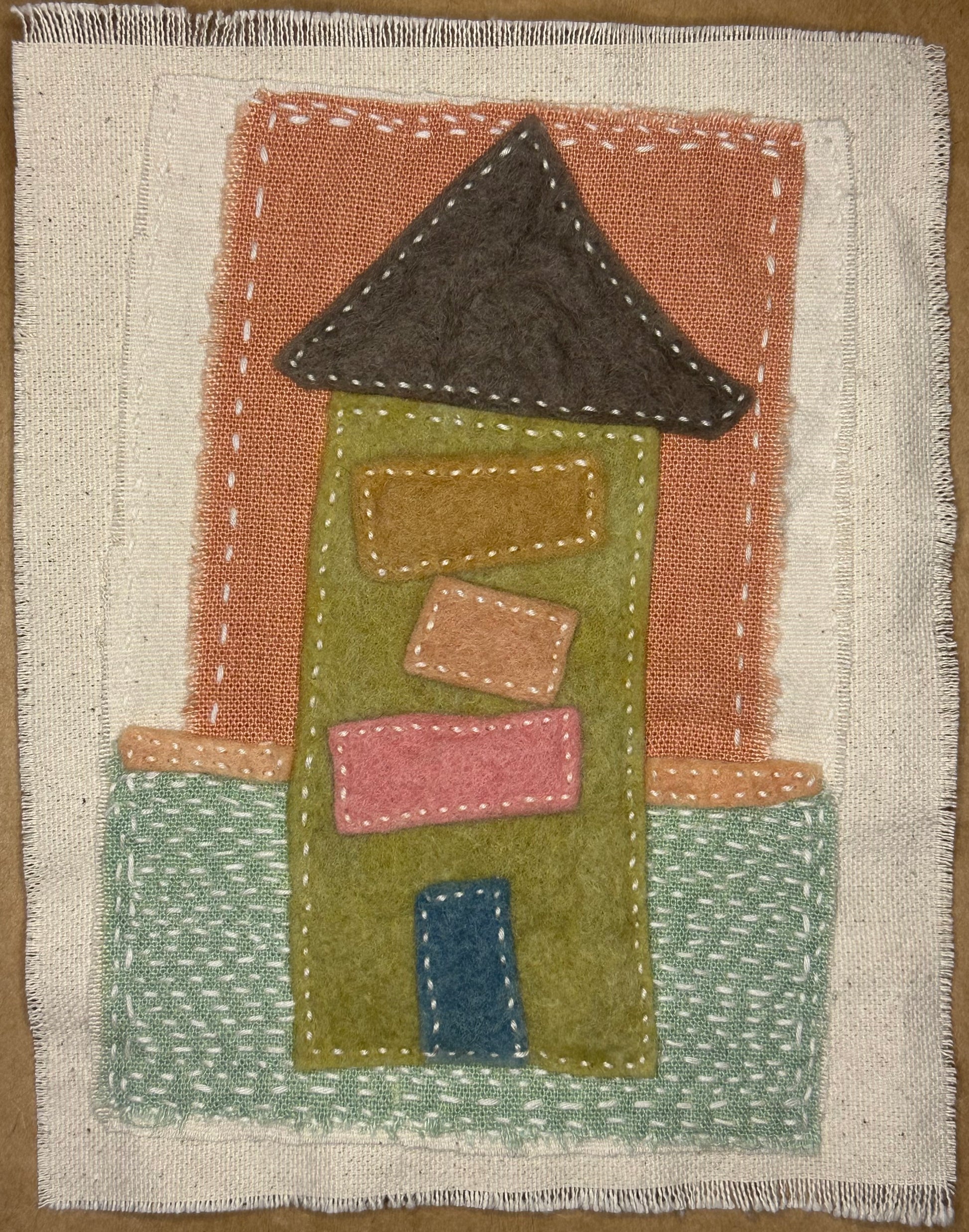 Fragments of Home textile art by Suzanne Gibbs with a green felt house, dark brown roof, pink and tan windows, blue door, and a base resembling mowed grass, hand-stitched in white.