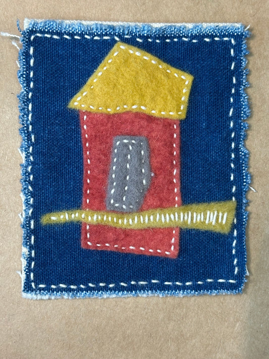 A small piece of artwork by Suzanne Gibbs featuring a collage of a rust-colored bird house with a blue sky, made from cotton, felt, and thread.