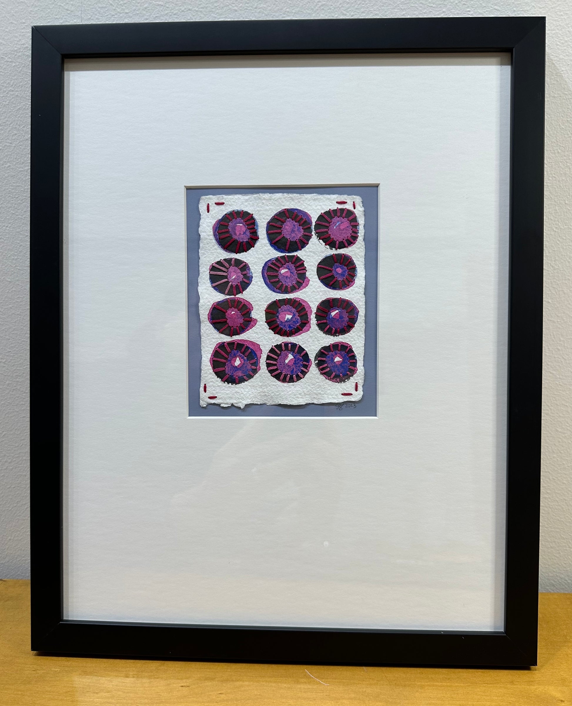 A framed artwork featuring a watercolor and flashe paint collage with sashiko thread on paper, showcasing a series of purple and black circular patterns, signed and dated by Suzanne Gibbs on the front.