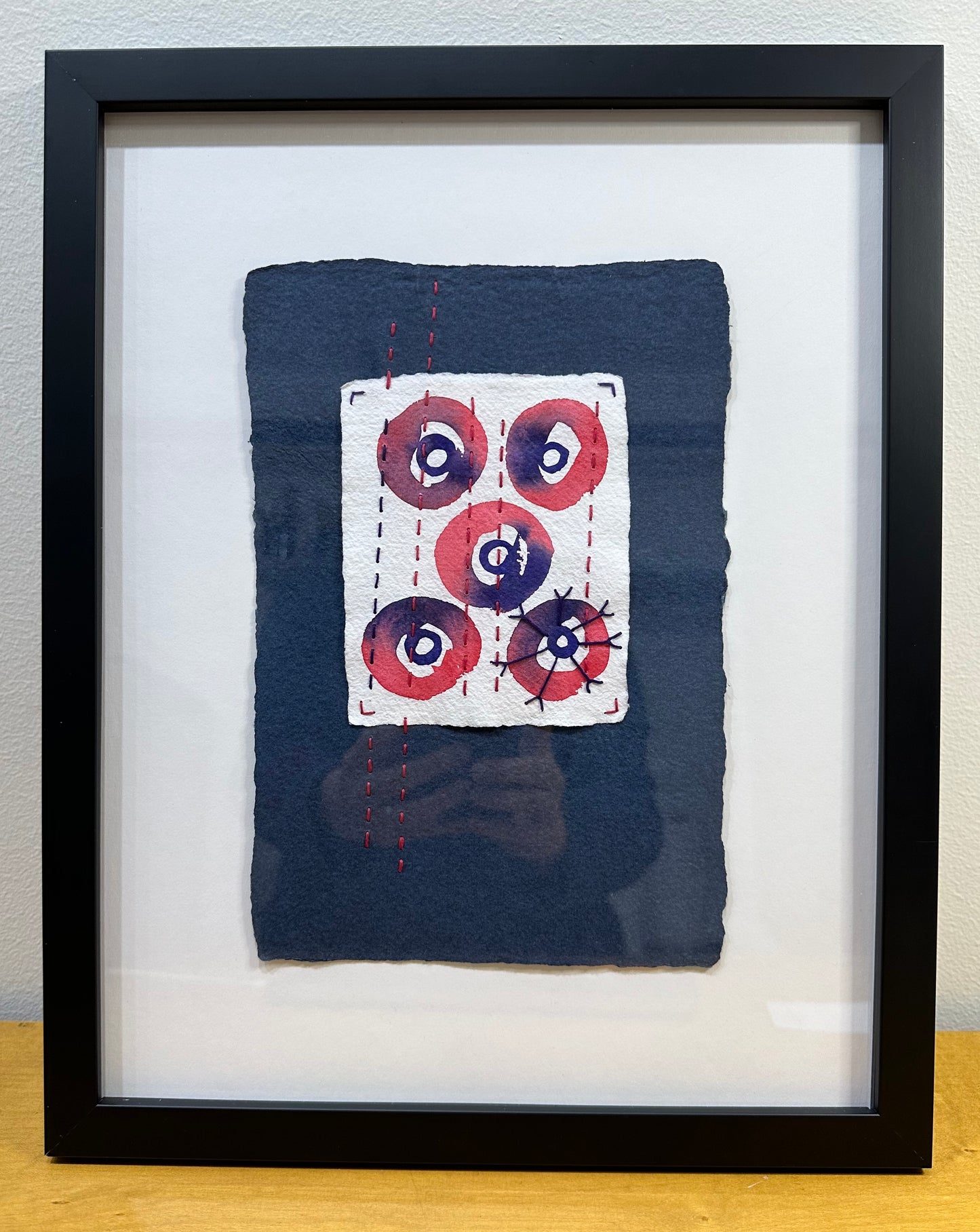 Mixed media artwork by Suzanne Gibbs from her Stitch & So series, created with watercolor paint and thread on handmade paper, featuring concentric circles with stitching over a deep blue background.